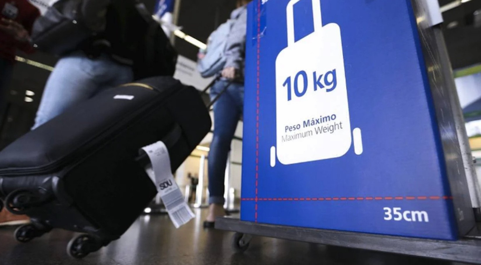 Pessoa puxa mala de rodinhas próxima a medidor de bagagem com limite de 10 kg em aeroporto brasileiro.