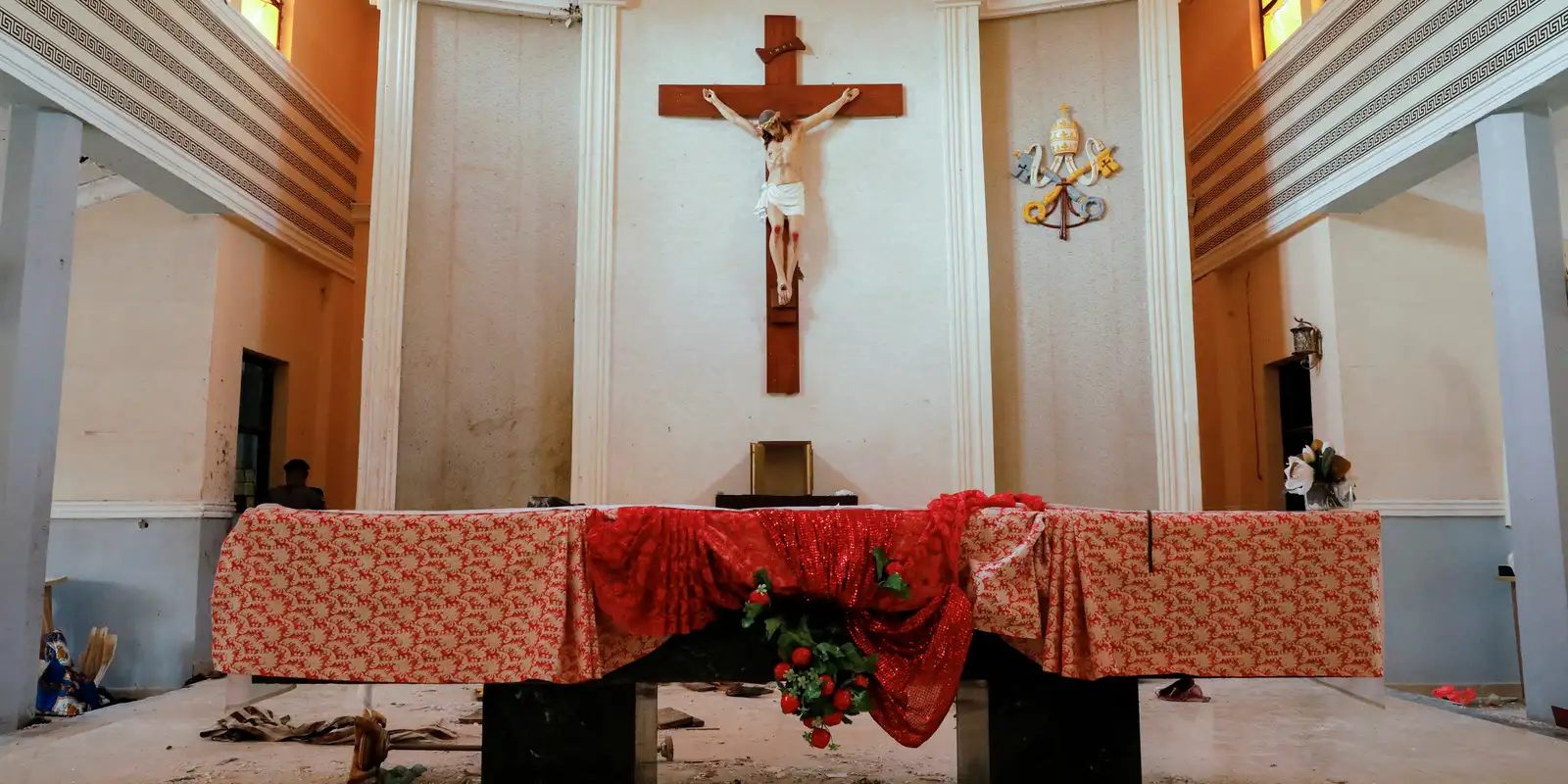 Interior de uma igreja com o altar parcialmente danificado, toalha vermelha caída e objetos fora do lugar, sugerindo sinais de ataque recente.