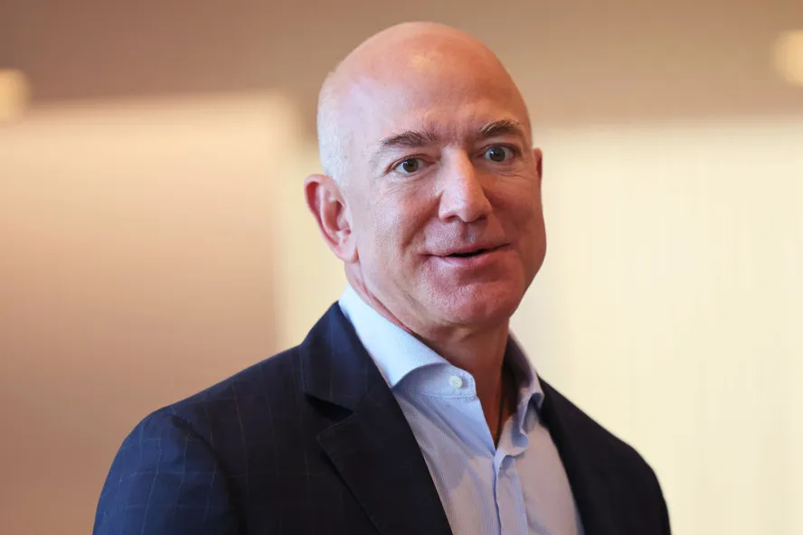 Jeff Bezos em ambiente interno, usando blazer azul escuro e camisa clara, olhando para a câmera com expressão neutra.