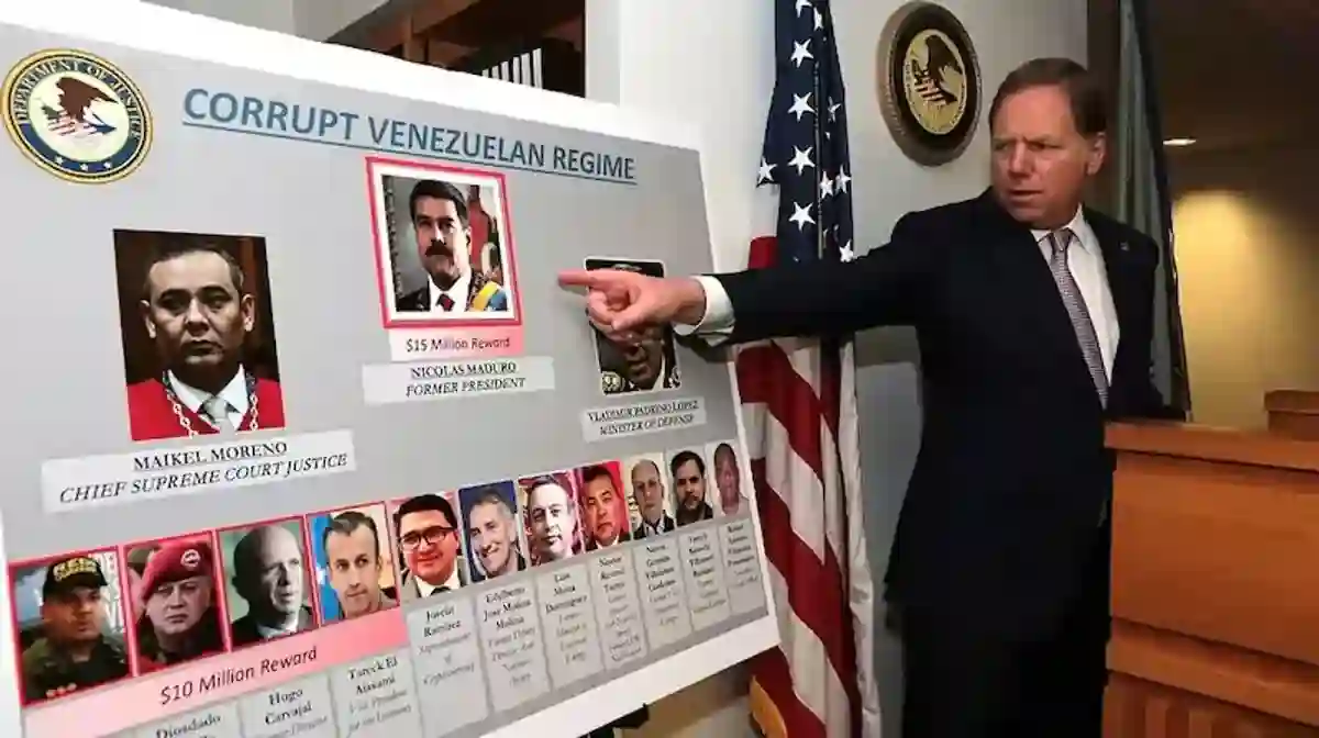 Oficial americano de terno escuro em coletiva de imprensa aponta para painel grande com título 'CORRUPT VENEZUELAN REGIME' exibindo fotos de Nicolás Maduro com recompensa de 15 milhões de dólares identificado como ex-presidente, Maikel Moreno como Chief Supreme Court Justice, fileira inferior com múltiplas fotos de oficiais venezuelanos com recompensas de 10 milhões de dólares, ao lado de bandeira americana, brasão do Departamento de Justiça dos EUA visível na parede