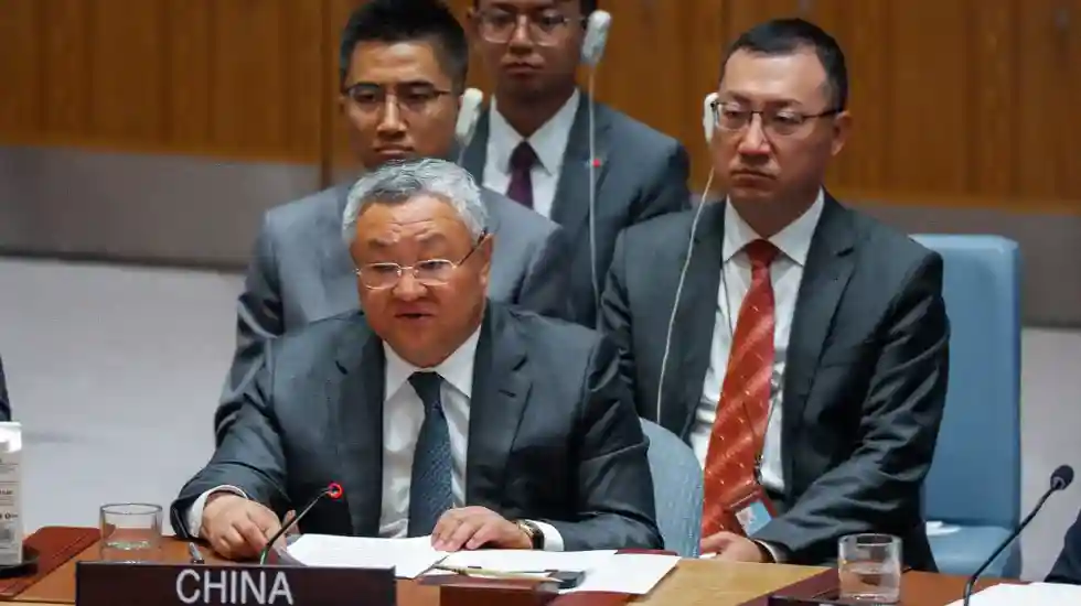 Embaixador da China na ONU Fu Cong sentado à frente em sessão do Conselho de Segurança, vestindo terno escuro e gravata azul, cabelos grisalhos, óculos, diante de placa identificadora 'CHINA', microfone com botão vermelho, copo de água e documentos sobre mesa, acompanhado por três diplomatas assessores ao fundo, dois de pé e um sentado usando gravata vermelha, em ambiente formal de reunião internacional