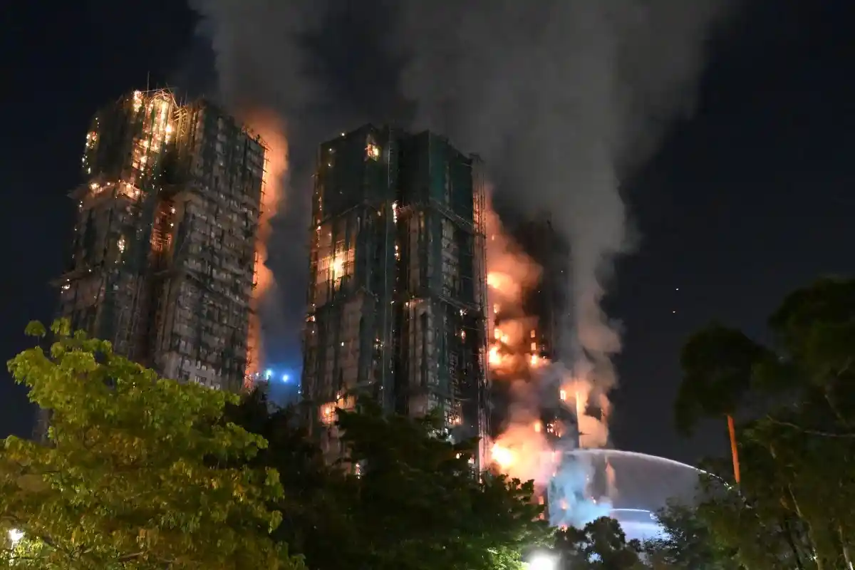 Três torres residenciais do complexo Wang Fuk Court em chamas durante a noite com fogo alaranjado nas fachadas e densa fumaça preta subindo enquanto bombeiros tentam controlar incêndio em Hong Kong