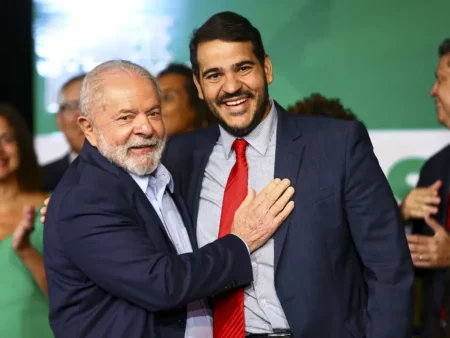 Dois homens de terno sorriem e posam abraçados em evento público, diante de um fundo verde desfocado com outras pessoas ao redor.