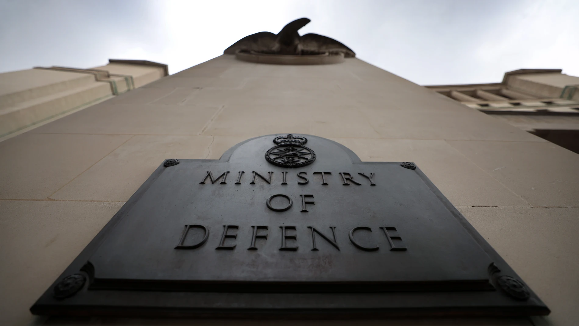 Placa metálica do Ministério da Defesa do Reino Unido fixada em prédio, fotografada de baixo para cima, com céu nublado ao fundo.