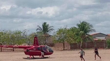 Helicóptero vermelho pousado em campo aberto enquanto três pessoas correm próximas ao local após a aterrissagem.