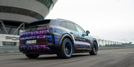 Porsche lança Cayenne elétrico com carregamento sem fio SUV elétrico da Porsche em teste de pista, visto por trás, com camuflagem colorida e rodas de aro azul em alta velocidade.