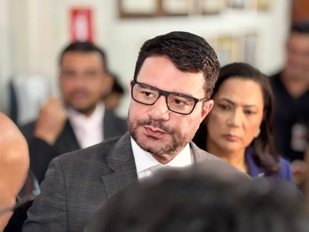 O governador Gladson Cameli fala com a imprensa, usando terno e óculos, enquanto pessoas ao fundo observam com expressão séria.