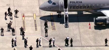 Agentes escoltam presos algemados na pista de aeroporto durante transferência, ao lado de avião estacionado.