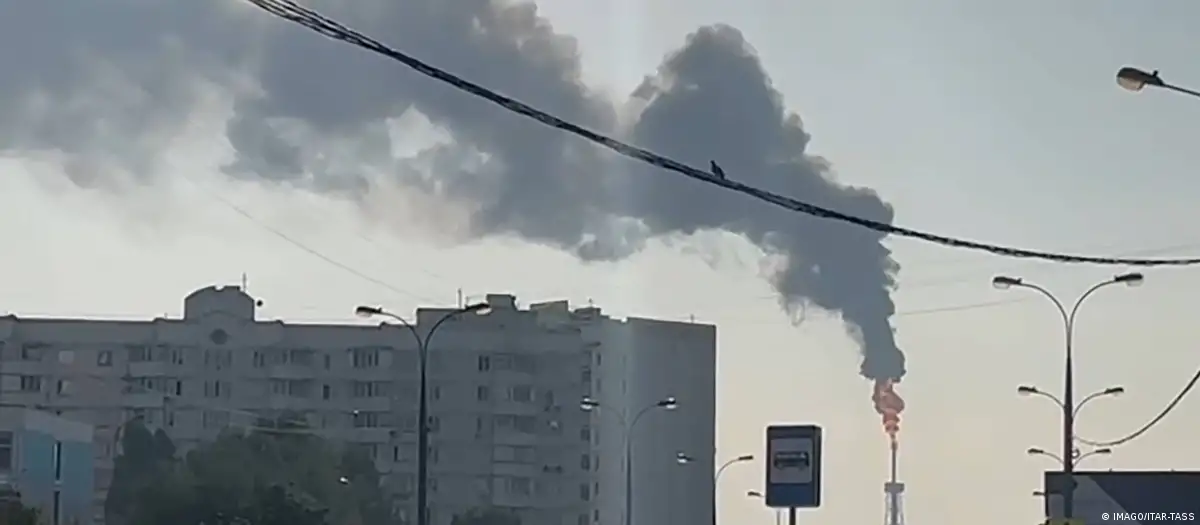 Coluna de fumaça negra subindo de uma usina atingida nos arredores de Moscou, com prédios residenciais ao fundo e luz do fim da tarde.