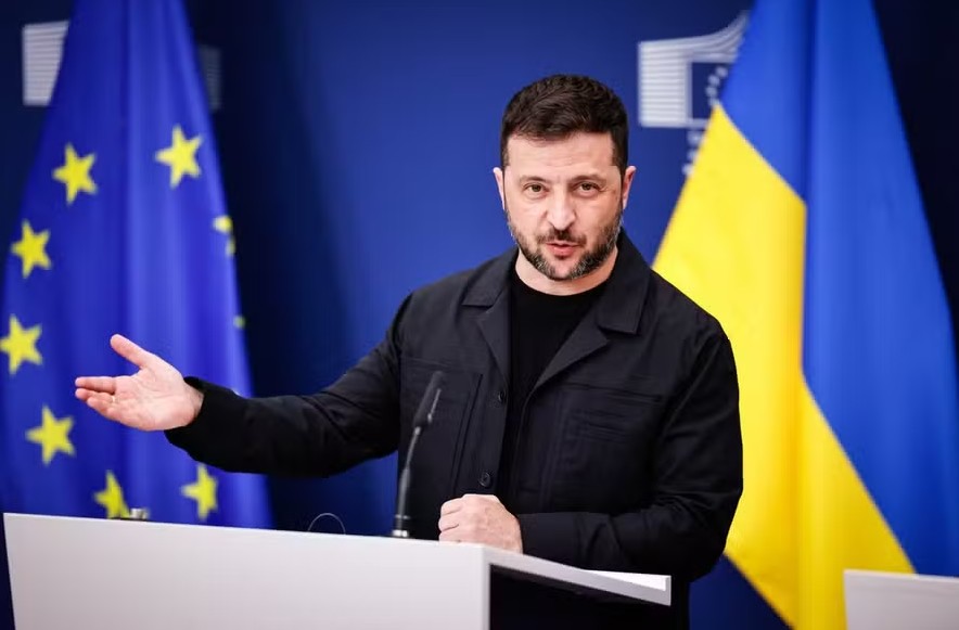 Homem em púlpito oficial gesticula durante pronunciamento, com bandeiras da União Europeia e da Ucrânia ao fund