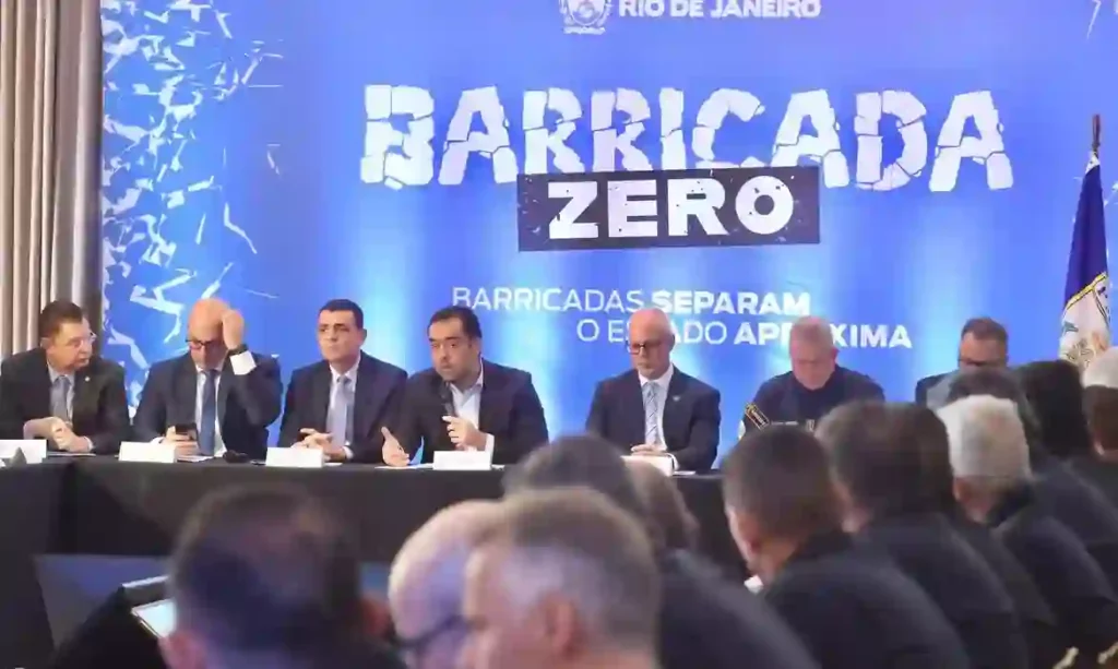 Rio inicia Operação Barricada Zero nas comunidades Coletiva de imprensa da Operação Barricada Zero com autoridades sentadas à mesa sob telão azul com logotipo do Rio de Janeiro e texto 'BARRICADA ZERO' em letras brancas, frase 'Barricadas separam o estado aproxima' visível, público de costas assistindo