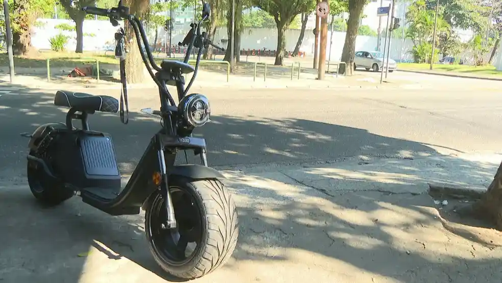 Ciclomotor elétrico estacionado em calçada de área urbana arborizada com rua asfaltada ao fundo em dia ensolarado