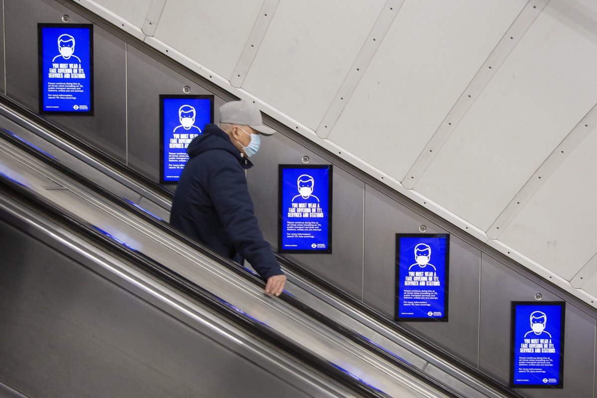 Homem usando máscara de proteção desce uma escada rolante em estação de metrô, ao lado de painéis azuis com avisos sobre obrigatoriedade de uso de máscara.