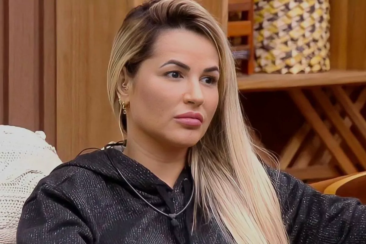 Deolane Bezerra sentada, olhando para o lado, vestindo blusa preta e com cabelo loiro solto.