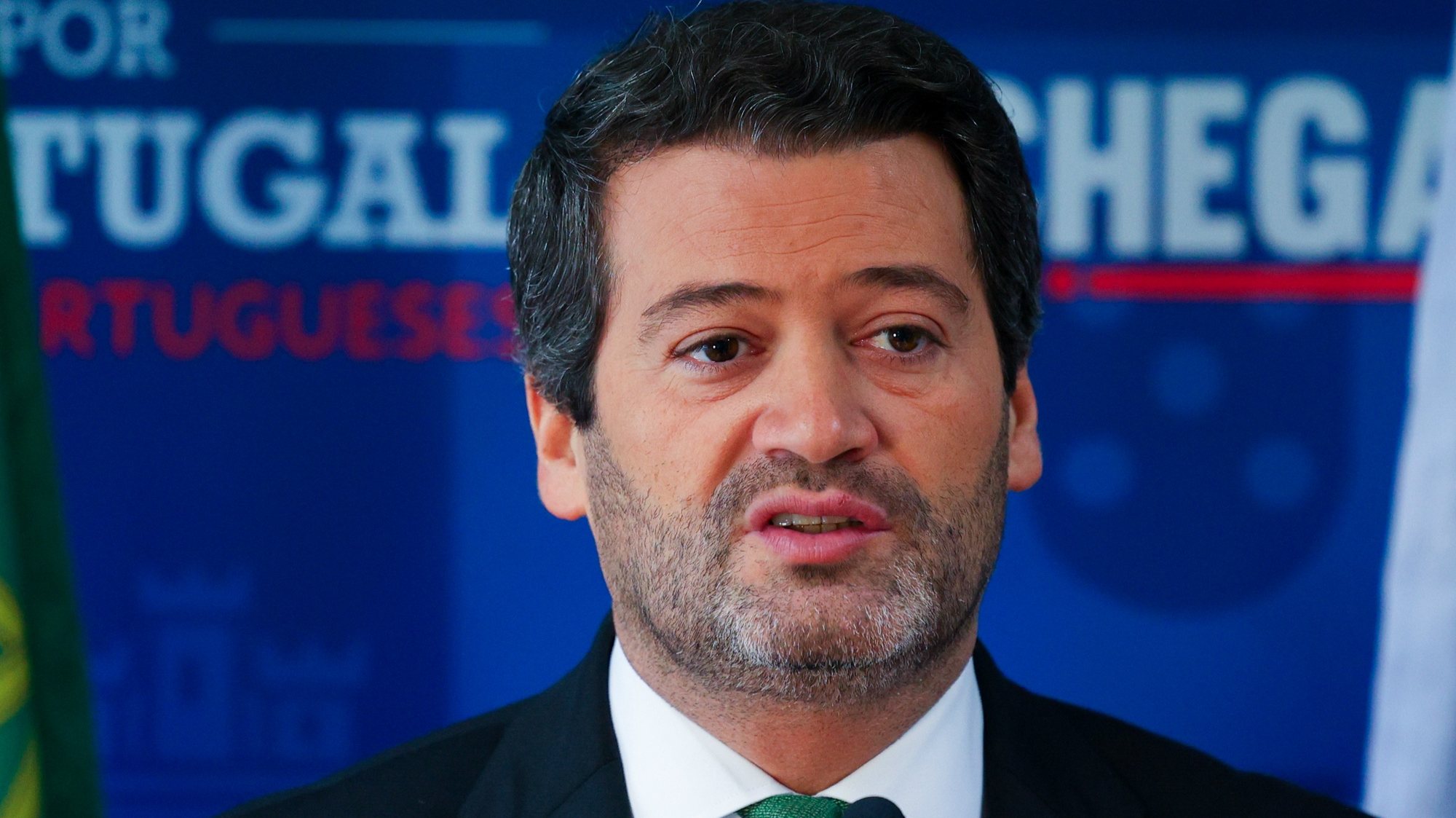 O deputado português André Ventura fala à imprensa durante entrevista em Lisboa, com fundo institucional do partido Chega ao fundo.