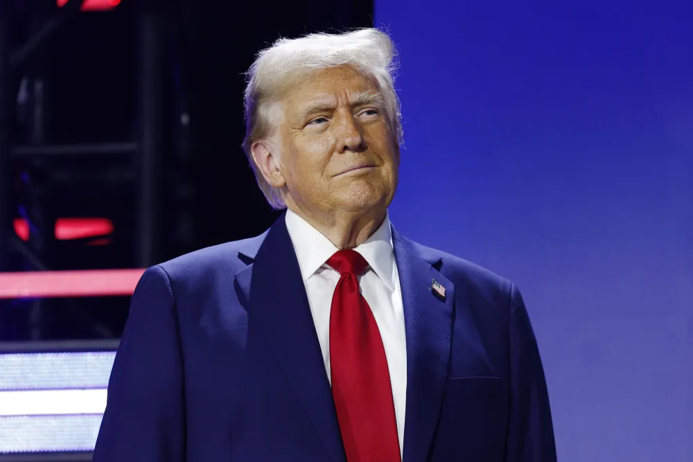 Donald Trump durante evento público, vestindo terno azul e gravata vermelha, sob iluminação azul, observando a plateia durante discurso político nos Estados Unidos.