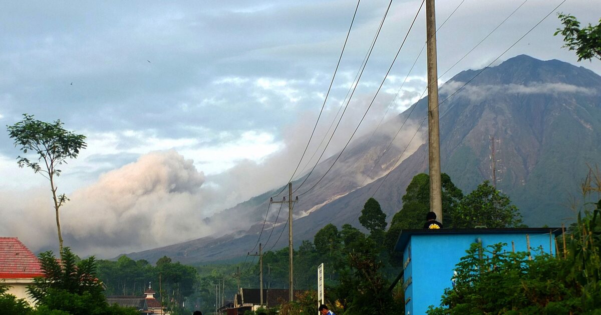 Nuvem de cinzas avança pela encosta de um vulcão em erupção, enquanto casas, vegetação e estruturas elétricas aparecem no primeiro plano da imagem.
