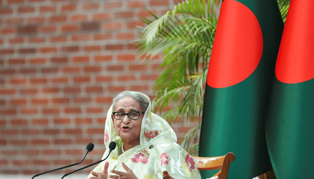 Sheikh Hasina fala ao microfone diante de duas bandeiras de Bangladesh, usando traje tradicional claro com estampa floral.
