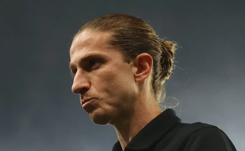 Homem de cabelo preso para trás observa o campo com expressão séria em ambiente esportivo.