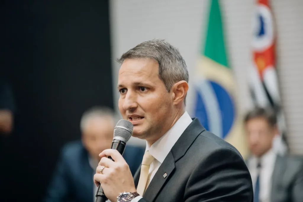 O secretário de Segurança Pública de São Paulo, Guilherme Derrite, fala ao microfone durante evento oficial, com bandeiras do Brasil e de São Paulo ao fundo.