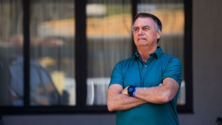Bolsonaro completa 100 dias de prisão domiciliar Homem de camisa verde posa de braços cruzados em frente a residência, sob luz natural, aparentando expressão séria.