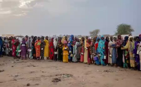 rupo de mulheres e crianças africanas vestindo roupas coloridas tradicionais aguardam em fila segurando recipientes metálicos em campo de refugiados localizado em região árida com tendas ao fundo