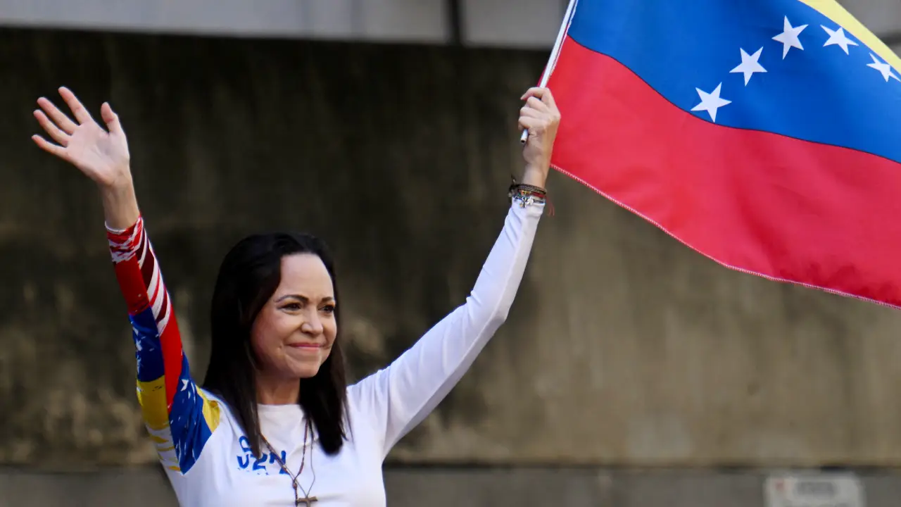 Maria Corina Machado segurando a bandeira da Venezuela com uma das mãos e acenando com a outra, em ambiente externo, durante ato público.