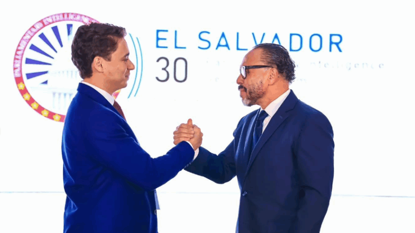 Dois homens em trajes formais cumprimentam-se com um aperto de mãos firme, em frente a um painel luminoso com elementos gráficos e o nome “El Salvador” ao fundo.