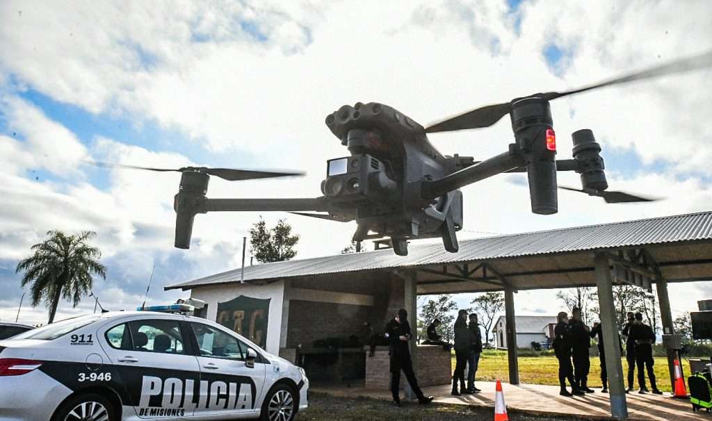 Drone de vigilância policial sobrevoando posto de controle fronteiriço argentino com viatura e agentes ao fundo