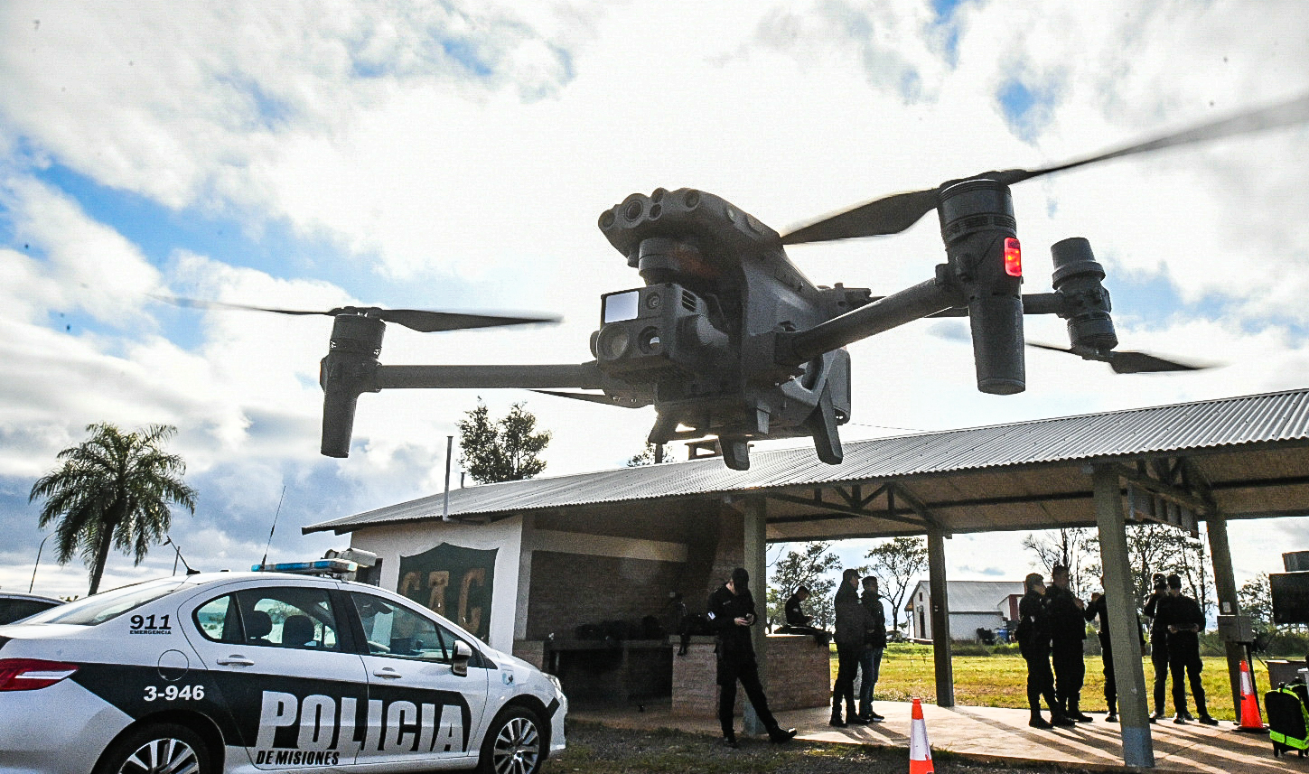 Drone de vigilância policial sobrevoando posto de controle fronteiriço argentino com viatura e agentes ao fundo