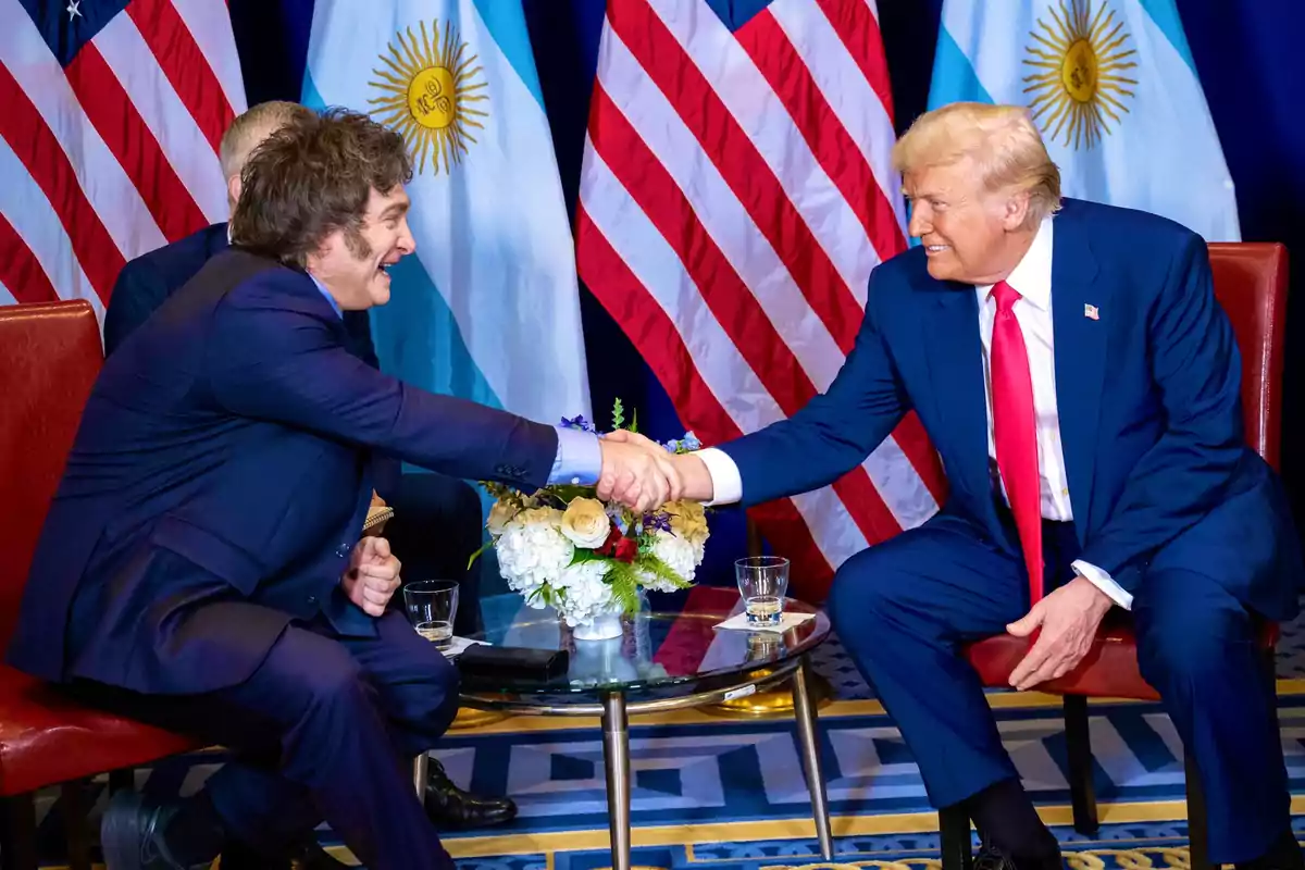 Dois líderes políticos sentados frente a frente apertam as mãos durante encontro diplomático, com bandeiras dos Estados Unidos e da Argentina ao fundo.