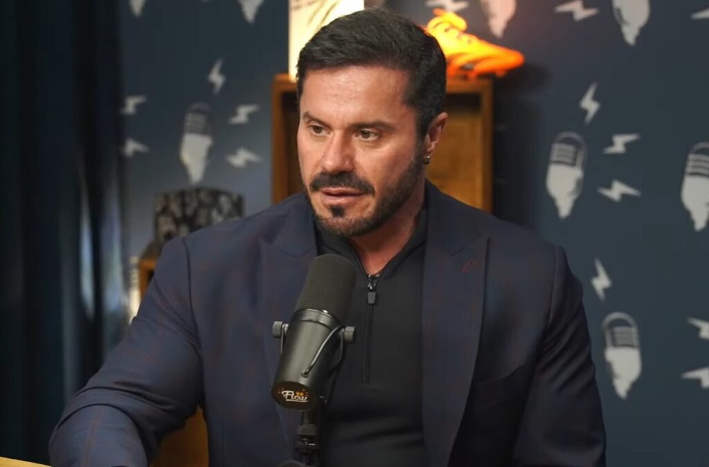 Homem de terno escuro fala em estúdio diante de microfone, durante entrevista gravada para programa de podcast.