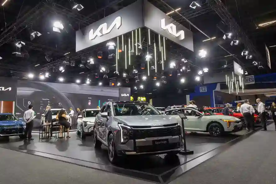 Estande da Kia no Salão do Automóvel de São Paulo 2025 com logotipo iluminado no teto, SUV Sportage prateado em destaque no centro sobre plataforma elevada, cercado por outros veículos da marca e visitantes circulando pelo pavilhão com iluminação profissional de holofotes