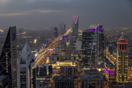 Vista aérea noturna da cidade de Riade, capital da Arábia Saudita, com arranha-céus iluminados e tráfego intenso.