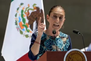 Presidente do México, Claudia Sheinbaum, discursando enfaticamente em um pódio com a bandeira nacional ao fundo, gesticulando com o dedo indicador levantado.