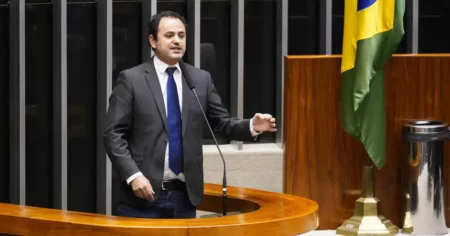 O deputado Glauber Braga, um homem de pele clara e cabelos escuros curtos, veste terno chumbo e gravata azul; ele está de pé na tribuna, gesticulando com a mão esquerda enquanto fala de forma expressiva ao microfone, com a bandeira do Brasil visível ao fundo.