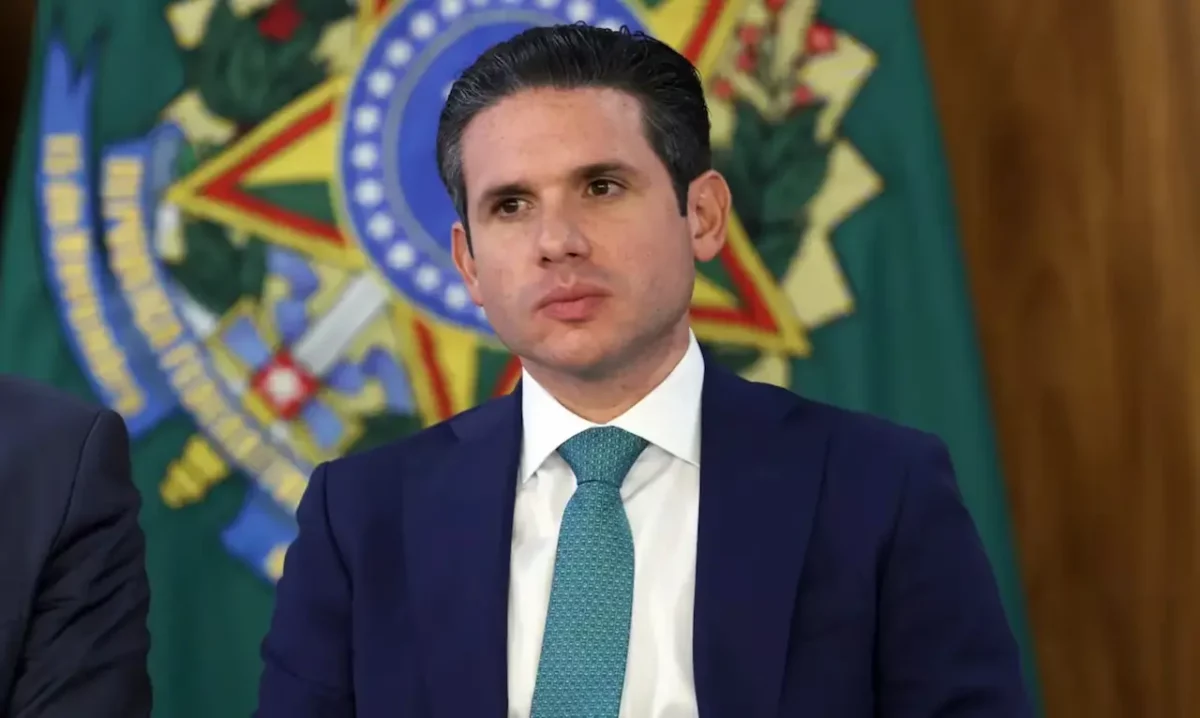 Hugo Motta, presidente da Câmara dos Deputados, aparece com expressão séria vestindo terno azul e gravata verde, posicionado à frente do Brasão da República durante sessão legislativa.