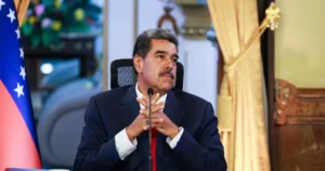 O presidente Nicolás Maduro, um homem de pele morena e bigode escuro, veste terno azul-marinho e gravata vermelha; ele está sentado com as mãos entrelaçadas à frente do corpo e olhar compenetrado, tendo a bandeira da Venezuela parcialmente visível ao fundo à esquerda.
