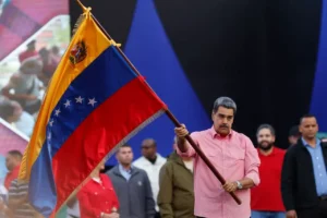 O presidente Nicolás Maduro, vestindo uma camisa social rosa de mangas compridas, segura com as duas mãos o mastro da bandeira da Venezuela, olhando para a frente com expressão séria; ao fundo, observam-se formas geométricas azuis e outras pessoas desfocadas.