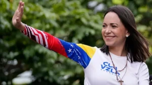 Maria Corina Machado sorrindo e acenando com a mão direita erguida, vestindo uma camisa branca com a sigla "Vzla" e cores da bandeira venezuelana na manga, além de um colar com crucifixo.