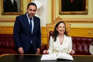 María Corina Machado, vestindo um blazer branco elegante, sorri abertamente enquanto está sentada assinando um livro de visitas oficial; ao seu lado, em pé e vestindo terno escuro, o presidente do Parlamento norueguês, Masud Gharahkhani, observa o momento com expressão cordial.