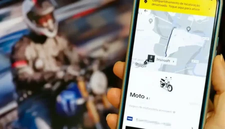 Em primeiro plano, uma mão segura um smartphone exibindo a interface de um aplicativo de corrida com a opção "Moto" selecionada na tela e um mapa de trajeto; ao fundo, fora de foco, vê-se um motociclista utilizando capacete e jaqueta pilotando uma moto.
