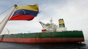 Um grande navio petroleiro de casco verde e vermelho navega em alto mar sob céu nublado; em primeiro plano, à esquerda, a bandeira da Venezuela tremula em um mastro, parcialmente focada.