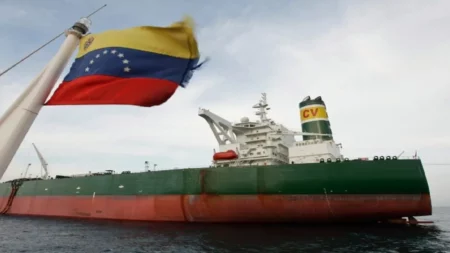 Um grande navio petroleiro de casco verde e vermelho navega em alto mar sob céu nublado; em primeiro plano, à esquerda, a bandeira da Venezuela tremula em um mastro, parcialmente focada.
