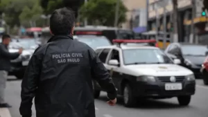 Policial civil de costas vestindo jaqueta preta com inscrição "Polícia Civil São Paulo" caminhando em rua movimentada perto de viatura.