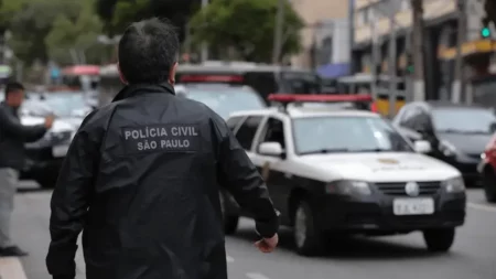 Policial civil de costas vestindo jaqueta preta com inscrição "Polícia Civil São Paulo" caminhando em rua movimentada perto de viatura.