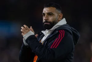 Mohamed Salah com expressão séria durante aquecimento, observado ao fundo pelo técnico Arne Slot.