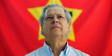 osé Dirceu, vestindo camisa social listrada de azul e branco, olha para o alto com expressão séria e compenetrada; ao fundo, preenchendo o cenário, vê-se a bandeira vermelha do Partido dos Trabalhadores com a estrela amarela centralizada.
